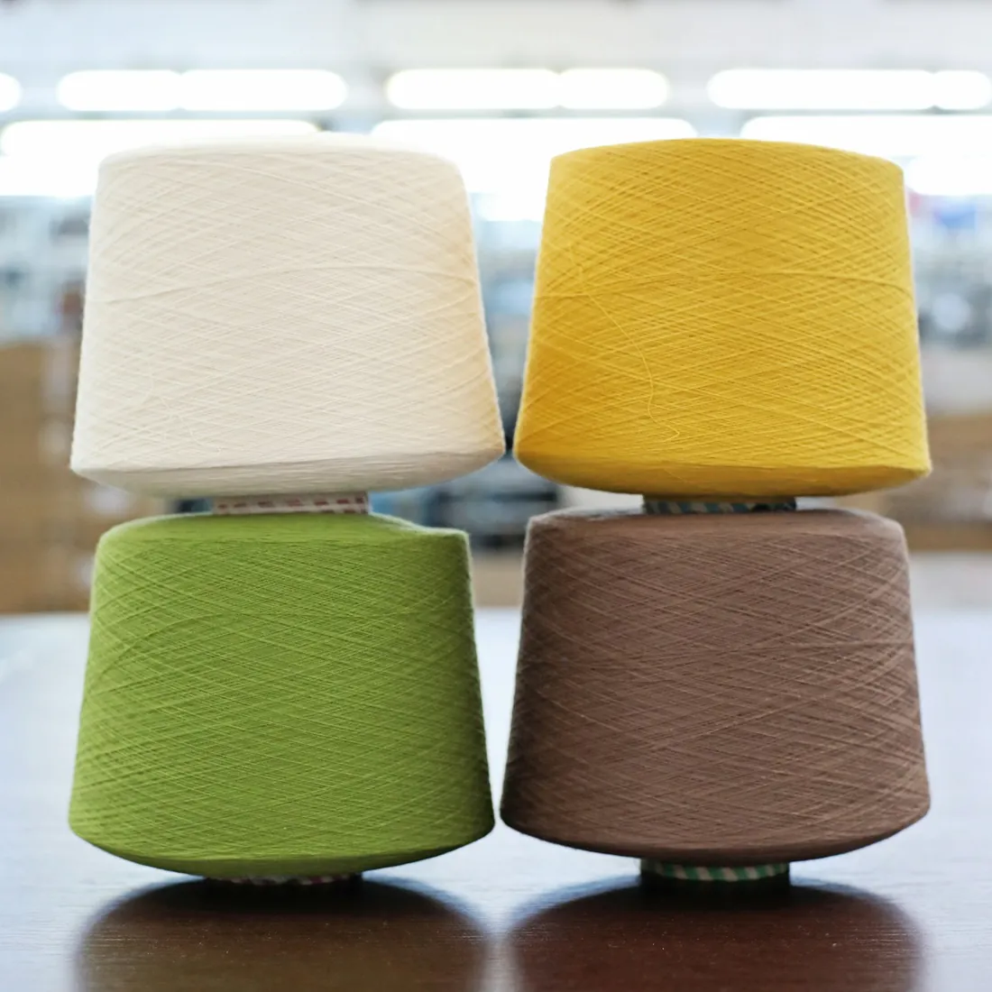 Conos de hilo de lana en color crudo, amarillo, verde y marrón apilados sobre una mesa de taller textil